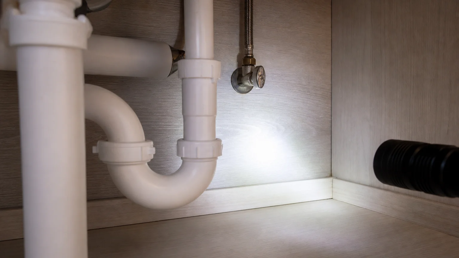 Flashlight beam sweeping the under-sink cabinet corner where roach droppings and smear marks collect first